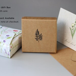 May include: A square brown kraft paper gift box featuring a black fern leaf stamp, positioned next to a small package wrapped in white floral paper and a cream-colored greeting card with a green flower illustration and the text Good Luck.