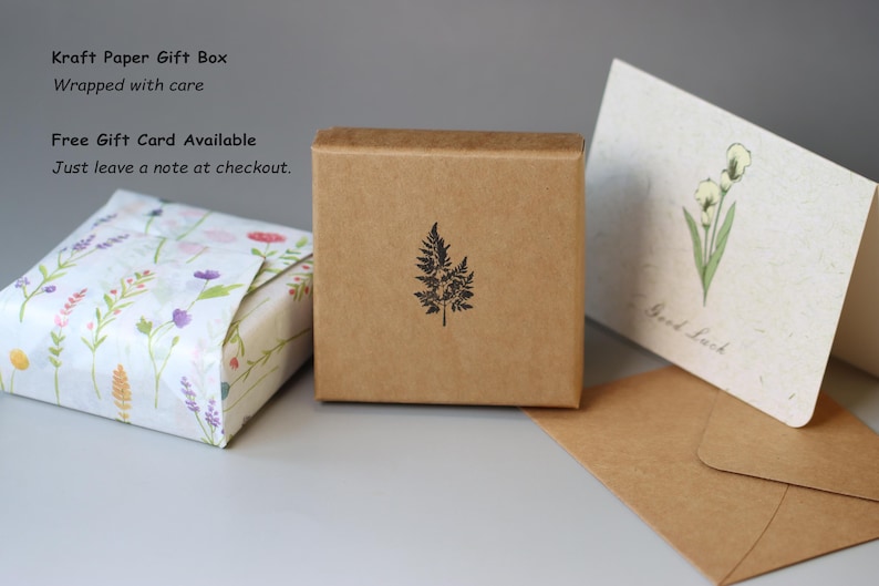 May include: A square brown kraft paper gift box featuring a black fern leaf stamp, positioned next to a small package wrapped in white floral paper and a cream-colored greeting card with a green flower illustration and the text Good Luck.