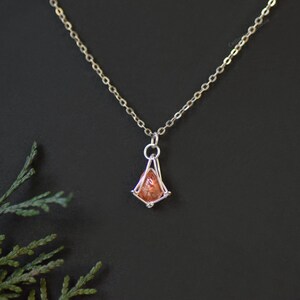 May include: A silver-toned metal chain necklace featuring a small, faceted, orange-hued gemstone pendant encased in a geometric wire-wrapped frame. The pendant hangs from a delicate link chain, showcasing a clear, warm-toned stone.