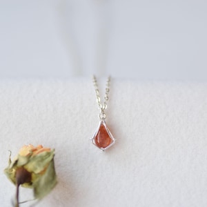 May include: A delicate silver necklace featuring a kite-shaped pendant with a translucent orange gemstone. The pendant is held by a thin silver wire frame and hangs from a fine silver chain, showcasing a minimalist and refined design.