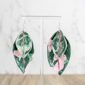 May include: A pair of leaf-shaped earrings. Each earring features two overlapping leaves in shades of green and pink, with a silver hook. The earrings are displayed against a white background.