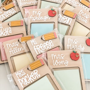 May include: A collection of wooden memo holders, each with a name in white script. Some feature a pencil or apple design. The holders contain colorful sticky notes and are packaged in clear plastic.