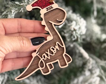 May include: A wooden dinosaur ornament featuring a dark brown wood grain body with a light wood border. The dinosaur wears a red glitter Santa hat with the year 2026 printed on the brim and the name Jaxon in script across the body.