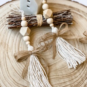 May include: Two white tassel garlands with brown burlap bows and wooden beads. The garlands are hanging from a small white ceramic house on a wooden slice.