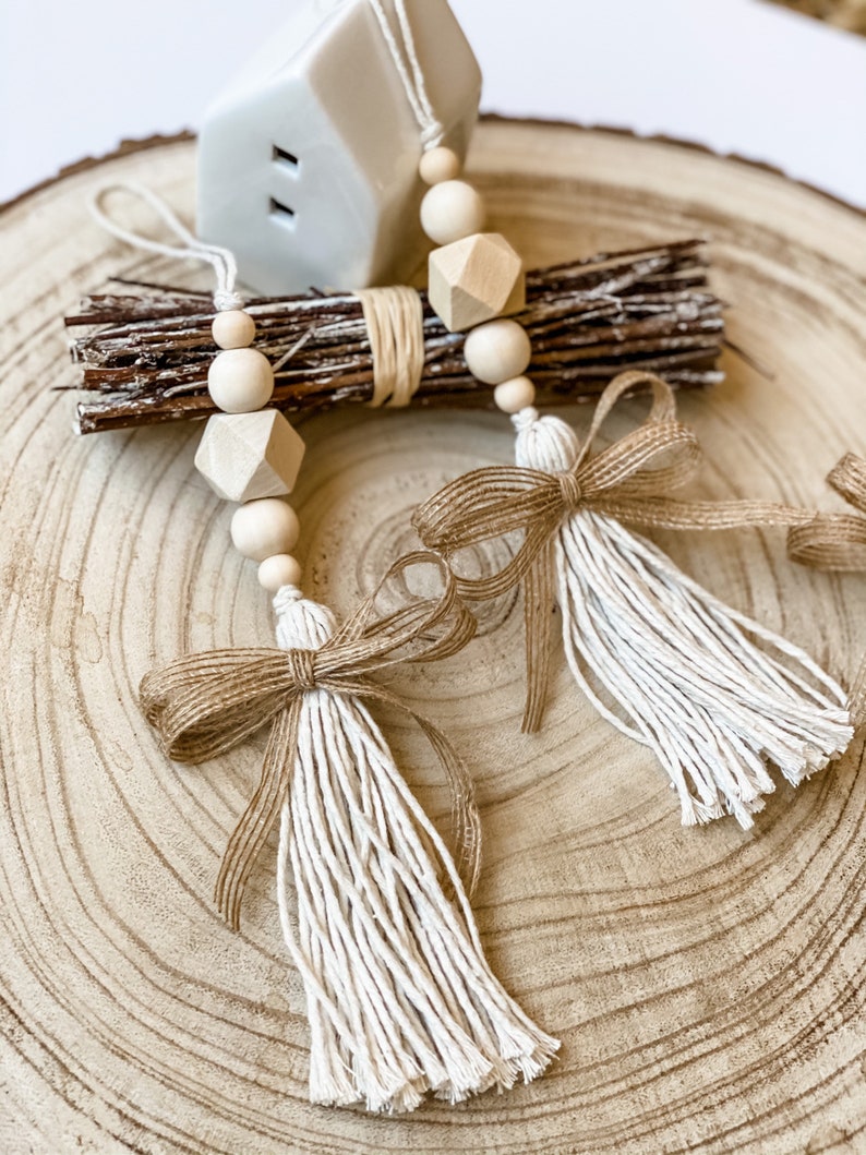 May include: Two white tassel garlands with brown burlap bows and wooden beads. The garlands are hanging from a small white ceramic house on a wooden slice.