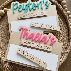 May include: Wooden sign with two sets of interchangeable signs for first and last day of school. The top set says "Peyton's" in teal and the bottom set says "Thalia's" in pink.