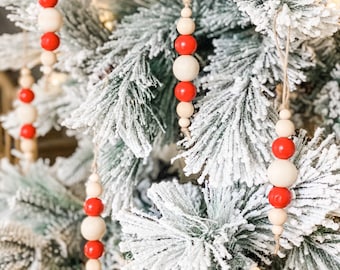 Op de afbeelding: Een close-up van een besneeuwde kerstboom versierd met rode en witte houten kralenversieringen. De ornamenten zijn aan een touwtje geregen en hangen aan de takken van de boom.