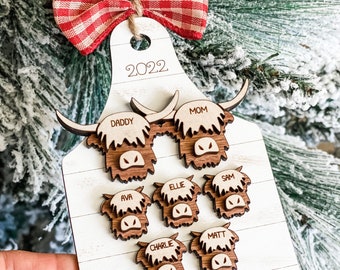 May include: A wooden Christmas ornament shaped like a cutting board with the year 2022 at the top. The ornament features a family of highland cow faces with names like "Daddy", "Mom", "Ellie", "Sam", "Ava", "Charlie", and "Matt". The ornament is decorated with a red and white checkered bow and the text "Our Herd".