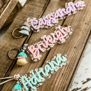 May include: Three personalized name keychains with daisy accents and tassel details. The names "Cassandra" in pink, "Breigh" in pink, and "Adriana" in light blue are featured. Each keychain has a gold-colored ring or a leather strap.