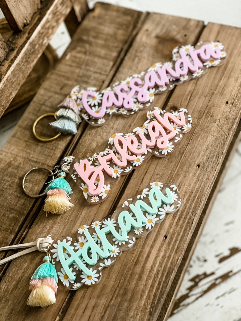 May include: Three personalized name keychains with daisy accents and tassel details. The names "Cassandra" in pink, "Breigh" in pink, and "Adriana" in light blue are featured. Each keychain has a gold-colored ring or a leather strap.