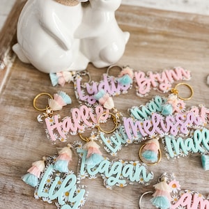 May include: A collection of colourful acrylic keychains with names written on them. Each keychain has a tassel and a daisy pattern. The names on the keychains are:  Lily, Reagan, Halle, Melissa, Lydia, Brynn, Sam, and Maile.