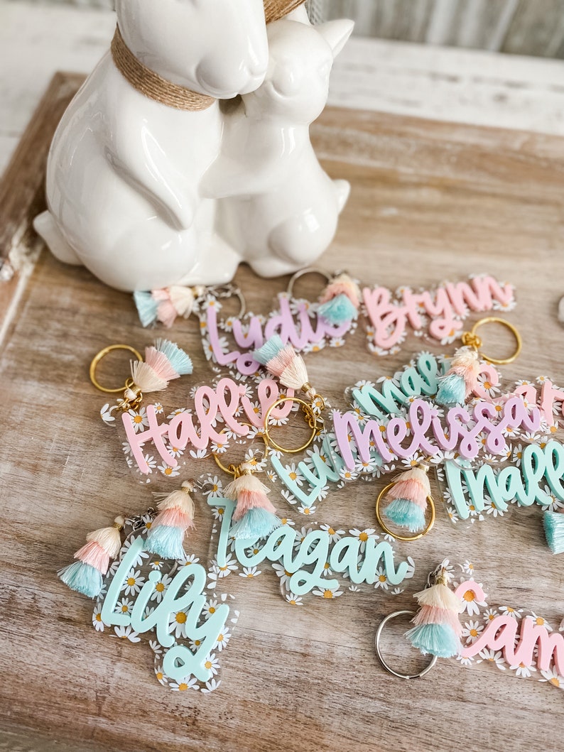 May include: A collection of colourful acrylic keychains with names written on them. Each keychain has a tassel and a daisy pattern. The names on the keychains are:  Lily, Reagan, Halle, Melissa, Lydia, Brynn, Sam, and Maile.