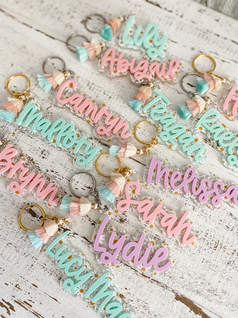 May include: A collection of personalised keychains with various names, including Lily, Abigail, Caitlyn, Mallory, Teagan, Reese, Bryn, Melissa, Sarah, Lydie, and Lucy Mae. Each keychain features a clear acrylic charm with a floral design and a colourful tassel.