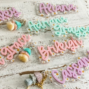 May include: Acrylic name keychains in various pastel colors, including pink, mint, and lavender. Each keychain features a name in a cursive font, set against a clear background with a daisy pattern. Some have tassel accents.