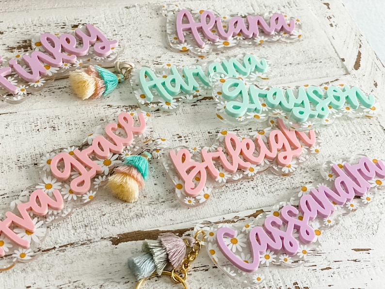 May include: Acrylic name keychains in various pastel colors, including pink, mint, and lavender. Each keychain features a name in a cursive font, set against a clear background with a daisy pattern. Some have tassel accents.