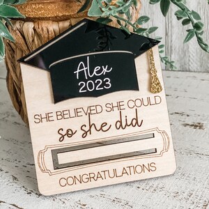 May include: Wooden graduation card with a black graduation cap cutout with the name Alex and the year 2023. The card reads "She believed she could so she did" with a cutout of a graduation scroll and the word "Congratulations".