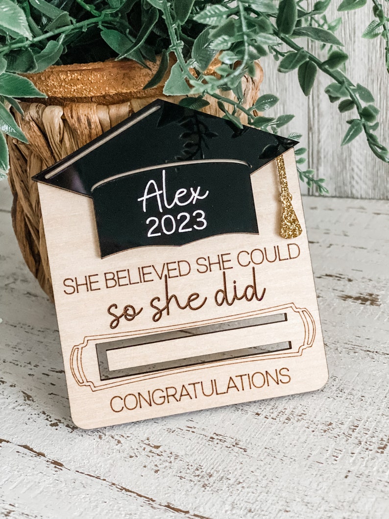 May include: Wooden graduation card with a black graduation cap cutout with the name Alex and the year 2023. The card reads "She believed she could so she did" with a cutout of a graduation scroll and the word "Congratulations".