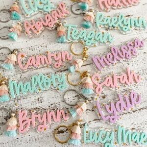May include: A collection of colourful acrylic keychains with daisy-patterned backgrounds and tassel charms. The keychains feature the names Lily, Abigail, Teagan, Camryn, Mallory, Brynn, Hallie, Raelynn, Melissa, Sarah, Lydie, and Lucy Mae.