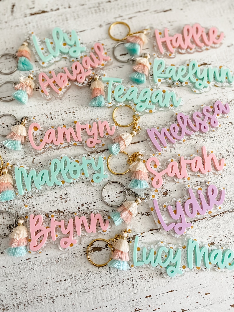 May include: A collection of colourful acrylic keychains with daisy-patterned backgrounds and tassel charms. The keychains feature the names Lily, Abigail, Teagan, Camryn, Mallory, Brynn, Hallie, Raelynn, Melissa, Sarah, Lydie, and Lucy Mae.