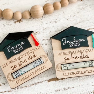 May include: Two wooden graduation money holders with a black graduation cap on top. The holders have a slot for a rolled-up bill. The text on the holders reads "Emma 2023 She believed she could so she did" and "Jackson 2023 He believed he could so he did".