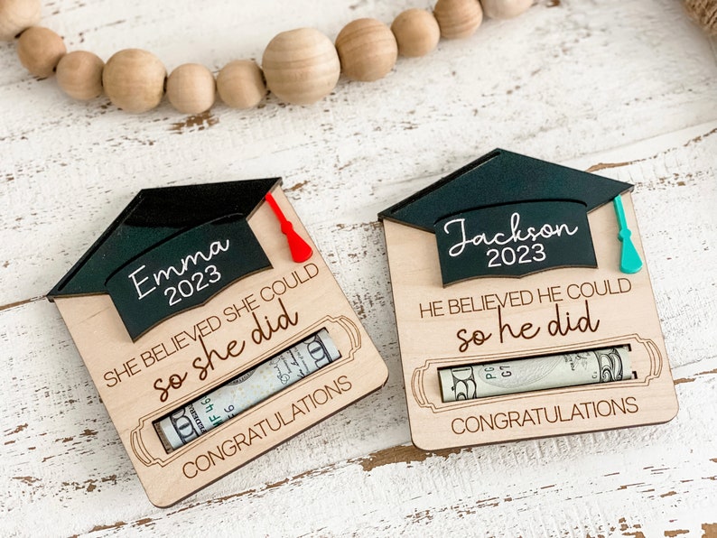 May include: Two wooden graduation money holders with a black graduation cap on top. The holders have a slot for a rolled-up bill. The text on the holders reads "Emma 2023 She believed she could so she did" and "Jackson 2023 He believed he could so he did".