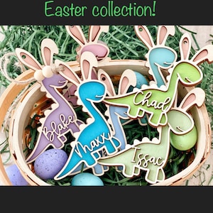May include: Five wooden dinosaur-shaped Easter decorations with bunny ears. The dinosaurs are painted in various pastel colours and have names written on them: Blake, Maxx, Chad, Issac. The decorations are in a basket filled with green grass.