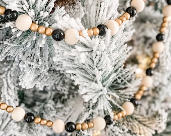 Könnte beinhalten: Ein weißer, flockiger Weihnachtsbaum, der mit einer Girlande aus schwarzen, weißen und goldenen Holzperlen geschmückt ist.