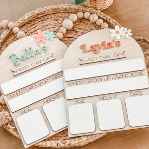 May include: Two arched wooden milestone signs with white dry-erase sections. One sign reads Bella's First Day Of, the other Layla's Last Day Of. Both feature When I Grow Up I Want To Be, Age, Height, and Date labels with floral accents at the top.