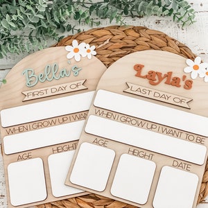 May include: Two arched wooden signs with white daisy accents. One sign features the text Bella's FIRST DAY OF, and the other features Layla's LAST DAY OF. Both include blank white rectangular spaces for writing age, height, date, and future career goals.