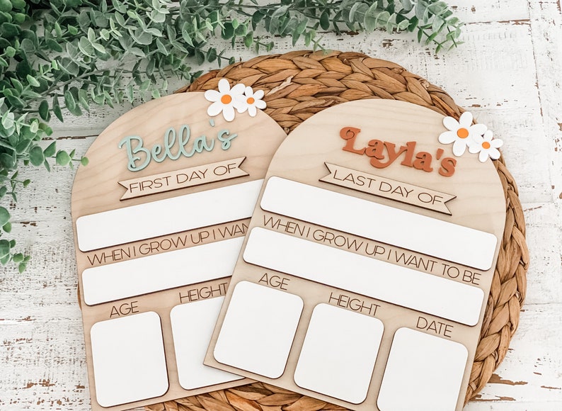 May include: Two arched wooden signs with white daisy accents. One sign features the text Bella's FIRST DAY OF, and the other features Layla's LAST DAY OF. Both include blank white rectangular spaces for writing age, height, date, and future career goals.