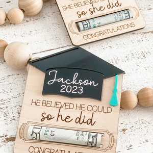 May include: A wooden graduation card with a black graduation cap cutout. The card has the text "Jackson 2023" and "He believed he could so he did". A rolled up dollar bill is visible in the slot on the card. The card also has the text "Congratulations".