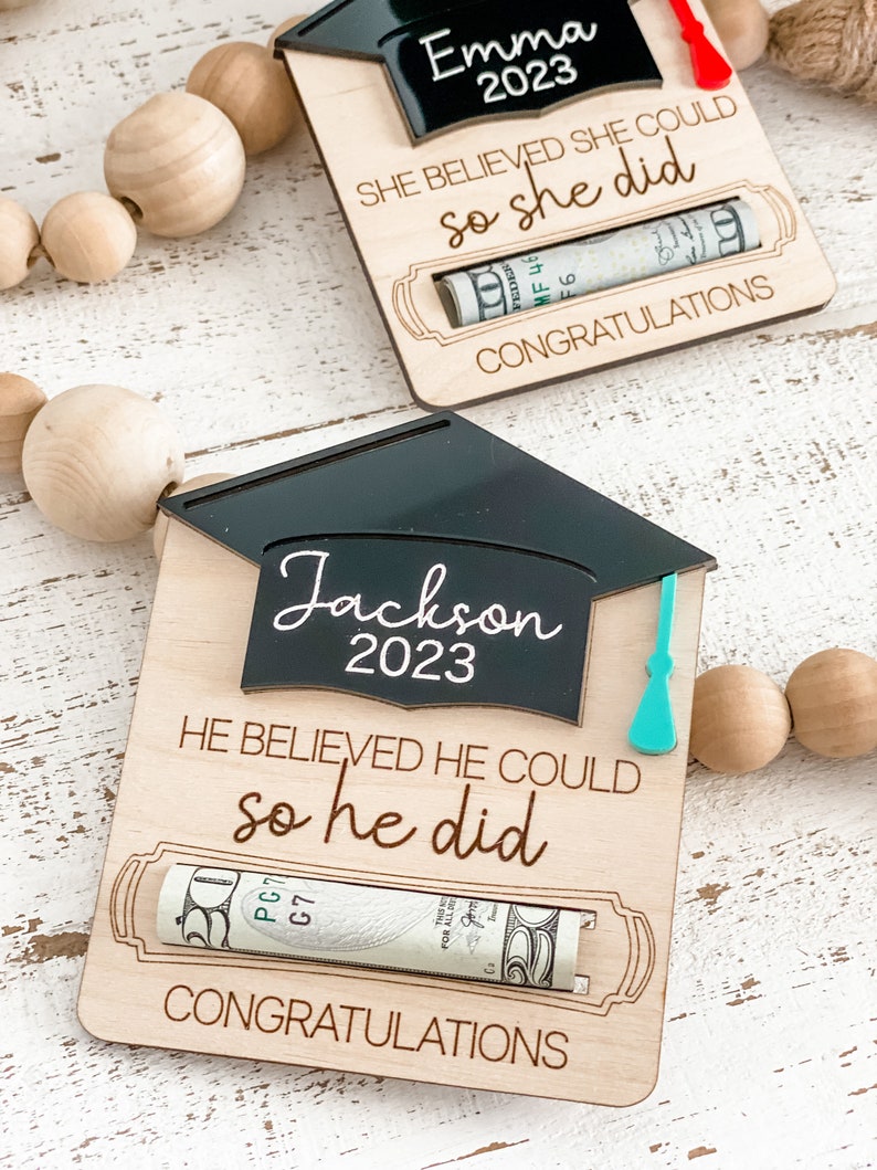 May include: A wooden graduation card with a black graduation cap cutout. The card has the text "Jackson 2023" and "He believed he could so he did". A rolled up dollar bill is visible in the slot on the card. The card also has the text "Congratulations".