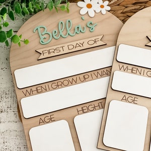 May include: An arched wooden sign featuring the text Bella's FIRST DAY OF, with white daisy accents. The board includes blank white spaces for writing information, with labels for WHEN I GROW UP I WANT TO BE, AGE, and HEIGHT in black text.