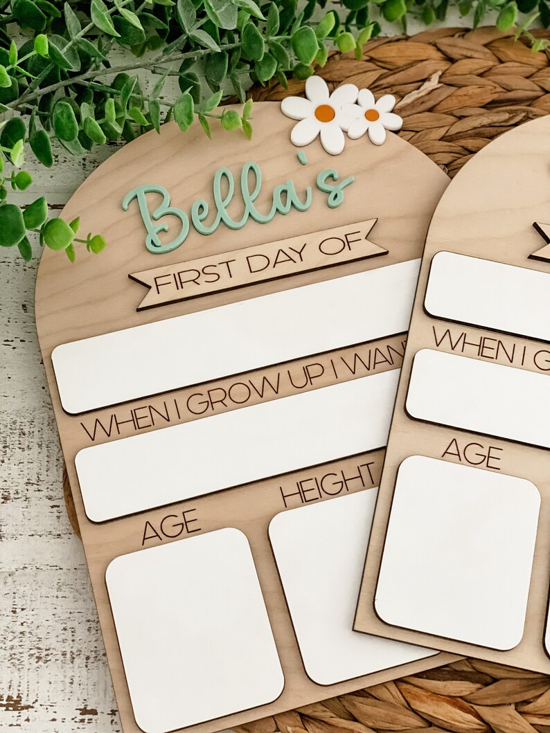 May include: An arched wooden sign featuring the text Bella's FIRST DAY OF, with white daisy accents. The board includes blank white spaces for writing information, with labels for WHEN I GROW UP I WANT TO BE, AGE, and HEIGHT in black text.