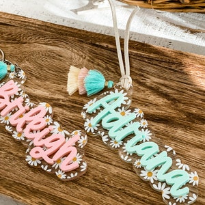 May include: Two personalized acrylic keychains with daisy accents. One keychain has the name "Brieigh" in pink, and the other has the name "Adriana" in mint green. Both keychains feature colorful tassels.