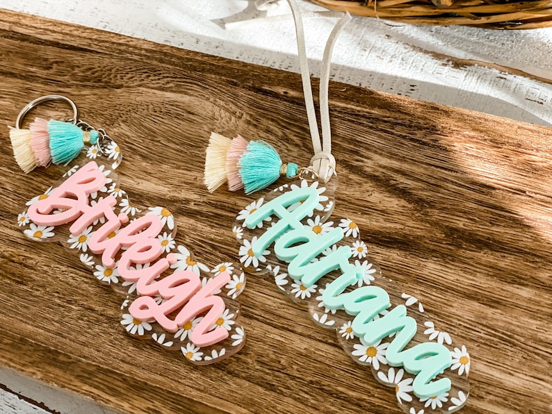 May include: Two personalized acrylic keychains with daisy accents. One keychain has the name "Brieigh" in pink, and the other has the name "Adriana" in mint green. Both keychains feature colorful tassels.
