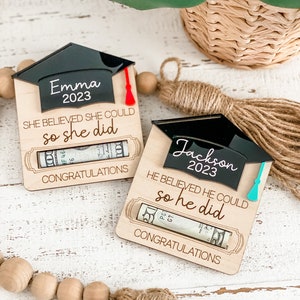 May include: Two wooden graduation cap-shaped boxes with a rolled-up dollar bill inside. The boxes are decorated with the names "Emma" and "Jackson" and the year "2023". The text "She believed she could so she did" and "He believed he could so he did" is printed on the boxes. The boxes are on a white wooden surface.