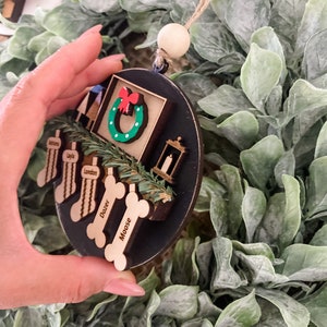 May include: A black and brown wooden Christmas ornament with a wreath, a lantern, and four dog bone-shaped tags with the names London, Doxer, and Moose. The ornament is hanging from a string with a wooden bead.