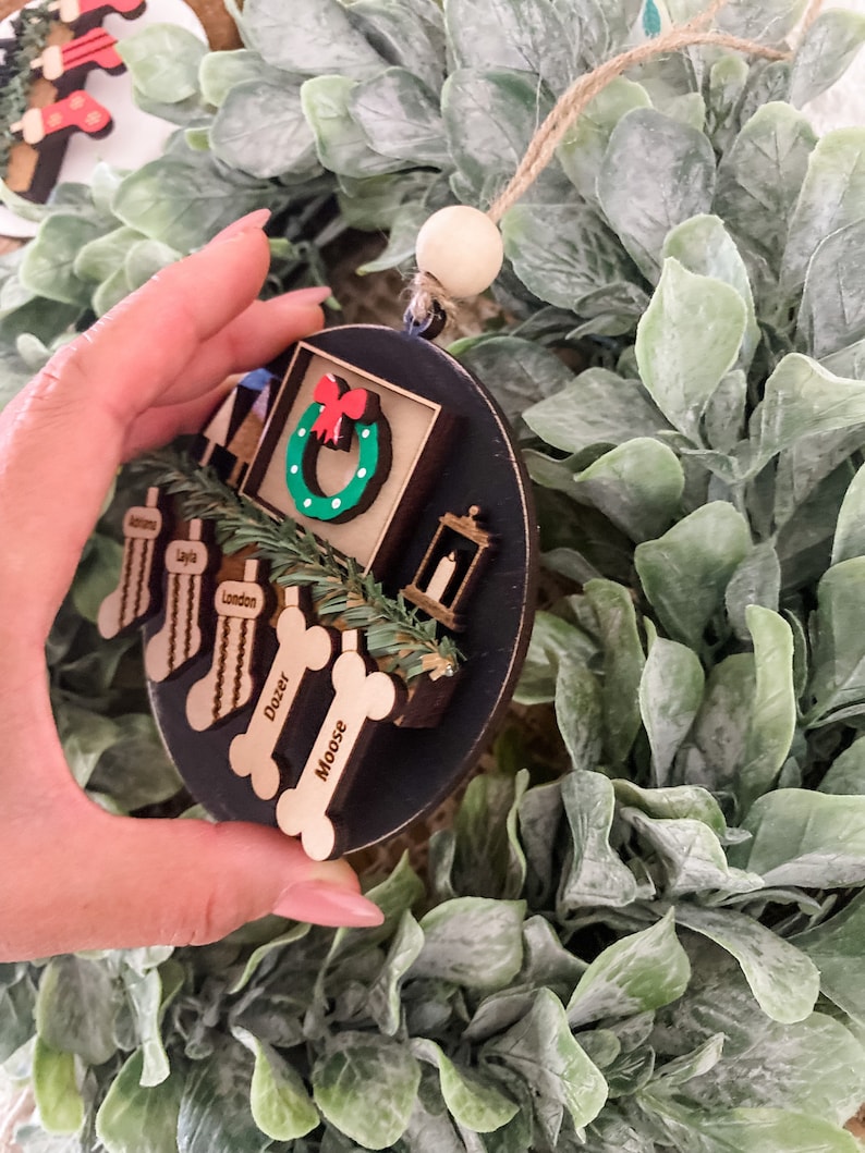 May include: A black and brown wooden Christmas ornament with a wreath, a lantern, and four dog bone-shaped tags with the names London, Doxer, and Moose. The ornament is hanging from a string with a wooden bead.