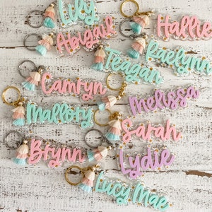 May include: A collection of 11 colourful acrylic keychain charms with daisy accents and tassel charms. The keychains feature the names: Lily, Abigail, Hallie, Raelynn, Teagan, Camryn, Mallory, Brynn, Melissa, Sarah, and Lydie Mae.