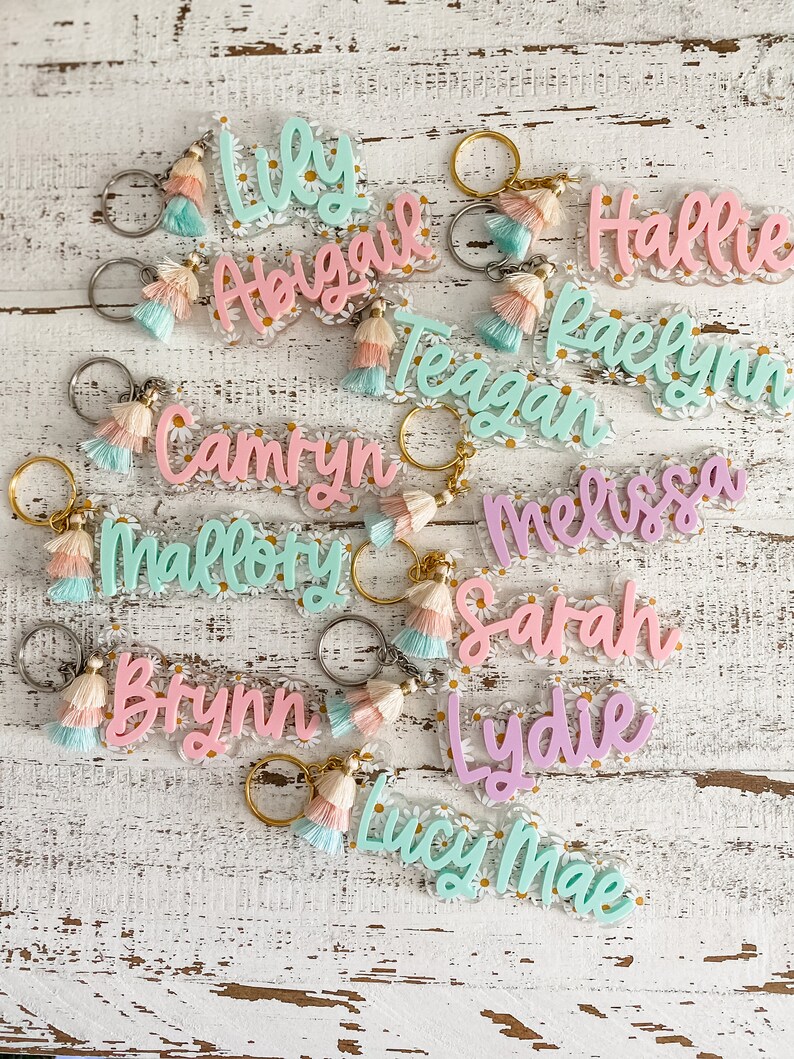 May include: A collection of 11 colourful acrylic keychain charms with daisy accents and tassel charms. The keychains feature the names: Lily, Abigail, Hallie, Raelynn, Teagan, Camryn, Mallory, Brynn, Melissa, Sarah, and Lydie Mae.