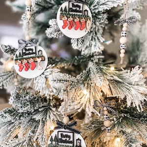 May include: Three round wooden Christmas ornaments with the text "Merry Christmas" and a shelf with stockings and names on each stocking. The ornaments are hanging on a flocked Christmas tree.