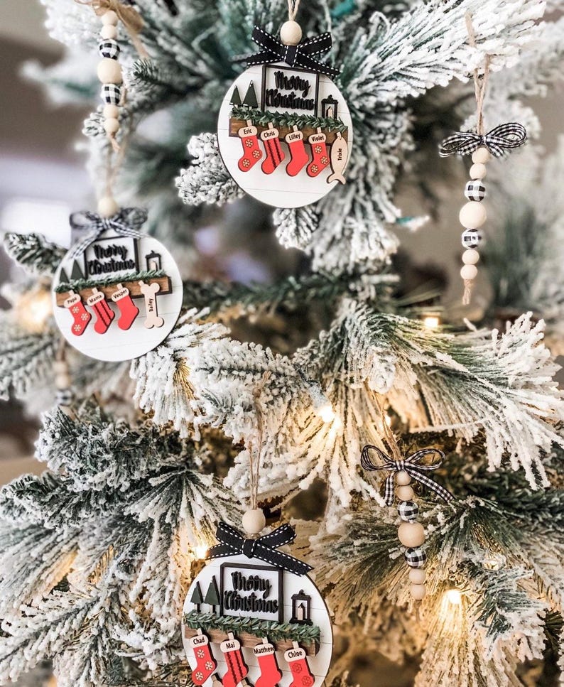 May include: Three round wooden Christmas ornaments with the text "Merry Christmas" and a shelf with stockings and names on each stocking. The ornaments are hanging on a flocked Christmas tree.