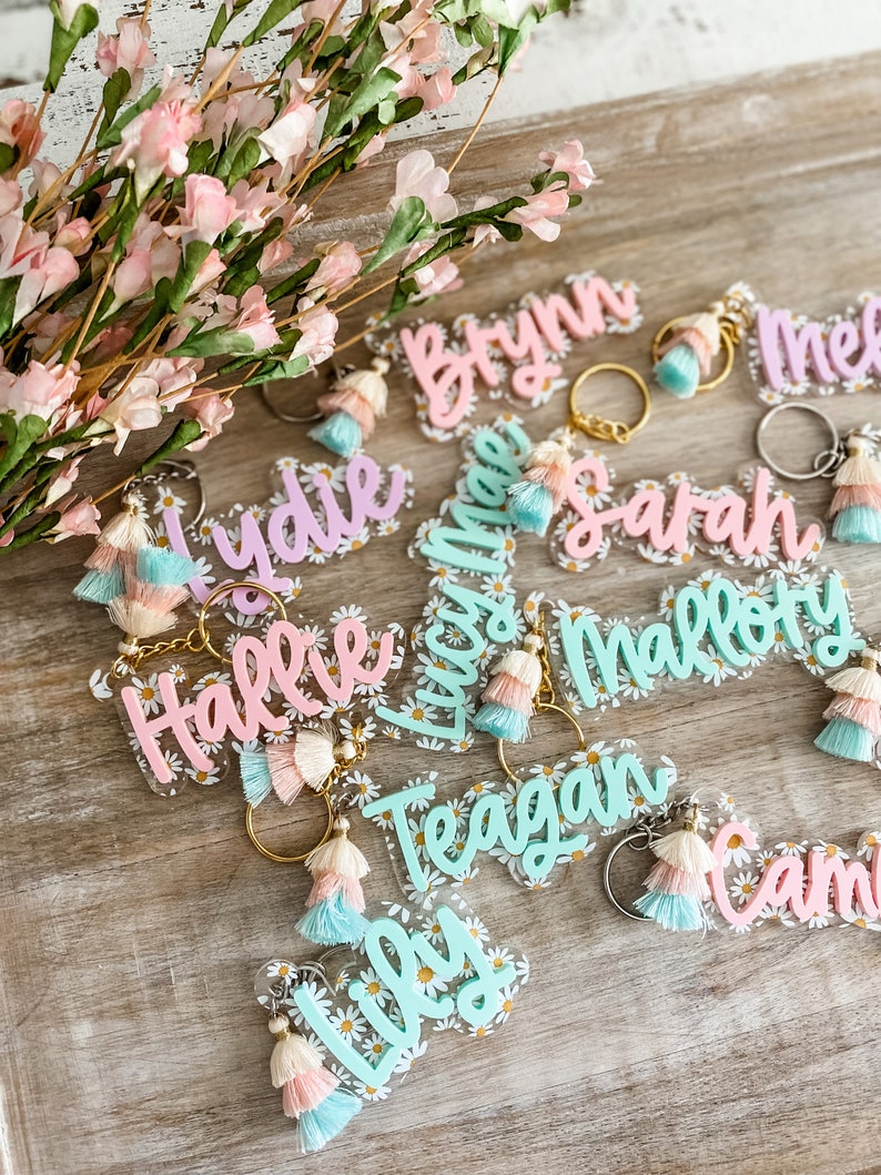 May include: A collection of colourful acrylic keychains with names written on them. Each keychain has a tassel and a daisy pattern. The names on the keychains are: Bryn, Mel, Lydie, Hallie, Lucas, Sarah, Mallory, Teagan, Lily, and Cami.