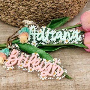 May include: Two daisy-themed acrylic name tags with tassels. One tag is light green with the name "Adriana" and the other is pink with the name "Braleigh". Both tags have a daisy border and are accented with colorful tassels.