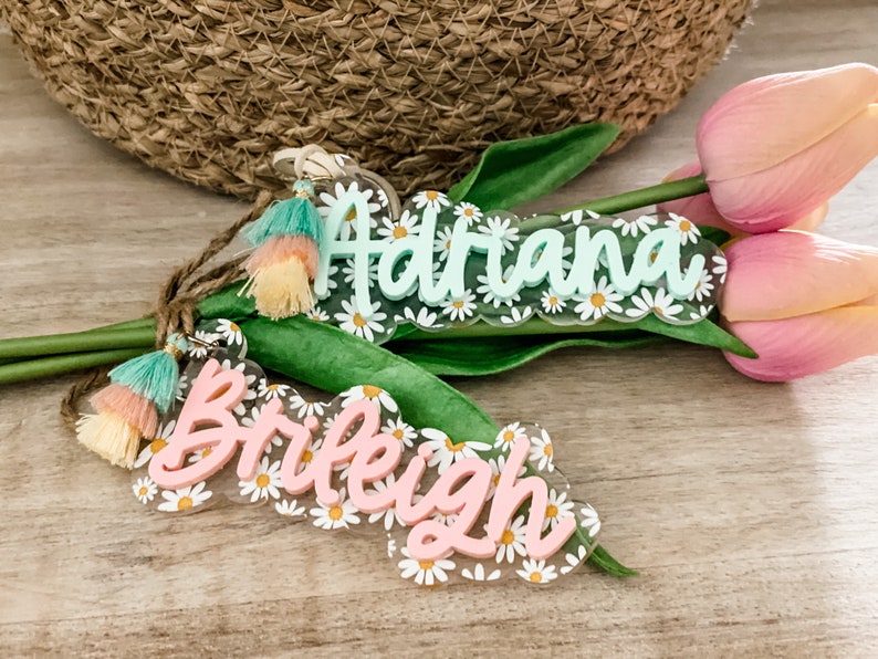 May include: Two daisy-themed acrylic name tags with tassels. One tag is light green with the name "Adriana" and the other is pink with the name "Braleigh". Both tags have a daisy border and are accented with colorful tassels.