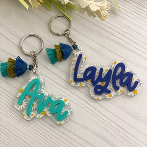 May include: Two personalized keychains. One says "Ava" in teal, the other says "Layla" in blue. Both names are on clear acrylic with a daisy pattern. Each keychain has a tassel with teal, navy, and olive green threads.