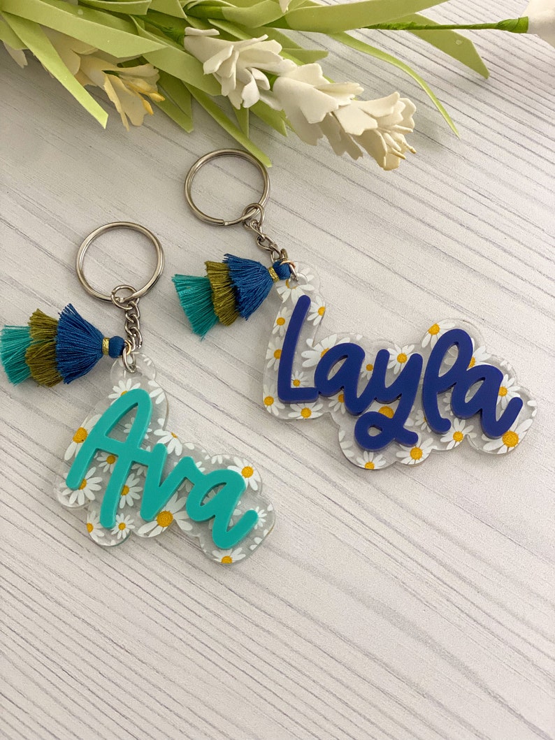 May include: Two personalized keychains. One says "Ava" in teal, the other says "Layla" in blue. Both names are on clear acrylic with a daisy pattern. Each keychain has a tassel with teal, navy, and olive green threads.