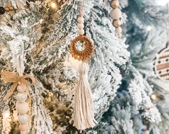 May include: A close-up of a Christmas tree decorated with a variety of ornaments, including a woven circle with a tassel, wooden beads, and a small bow. The tree is covered in artificial snow.