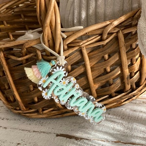 May include: A clear acrylic name tag with the name "Adriana" in a light green script, surrounded by white daisies. The tag has a scalloped edge and a tassel with pastel colors. The tag is hanging from a woven basket.